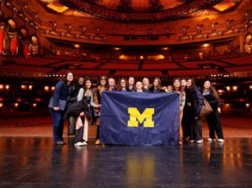 Students standing on the Fox Theater stage holding a UM flag, with the whole theater seating behind them.